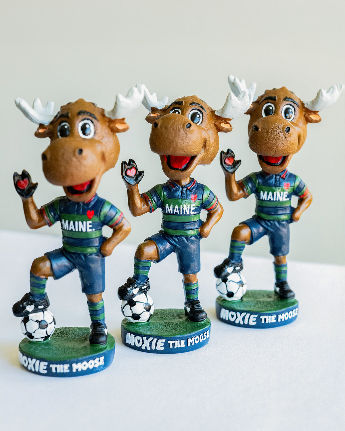 Three bobblehead figures of a moose mascot in Maine sports attire on a white background.