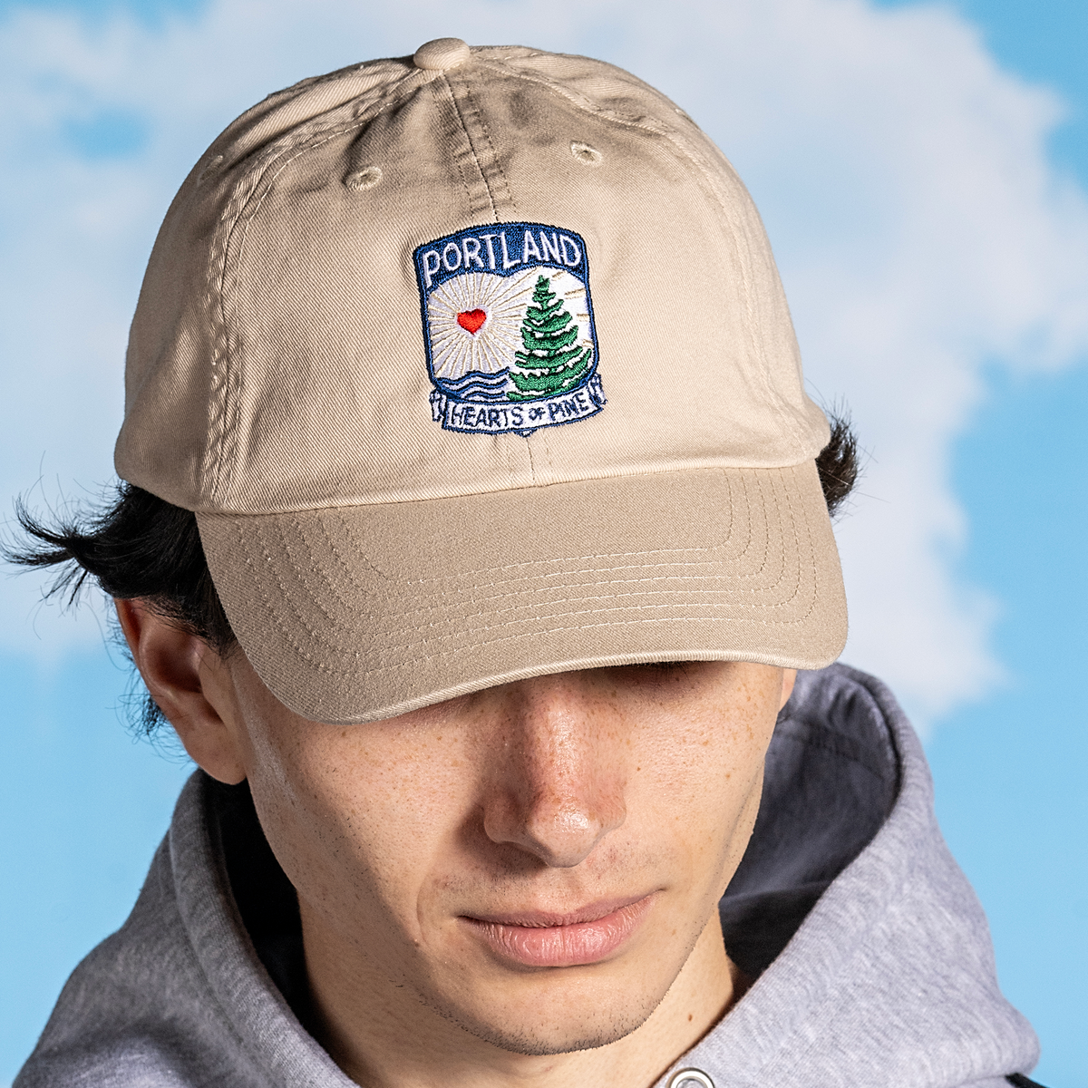 A person wearing a beige 'Hearts of Pine Dad Hat' with navy detailing and an embroidered patch on the front that reads 'PORTLAND Hearts of Pine'.