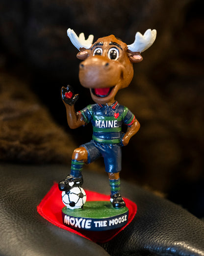 Bobblehead of a moose mascot wearing a Maine jersey with a soccer ball on a dark surface.