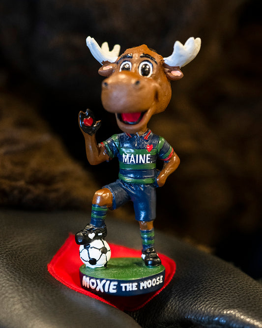 Bobblehead of a moose mascot wearing a Maine jersey with a soccer ball on a dark surface.