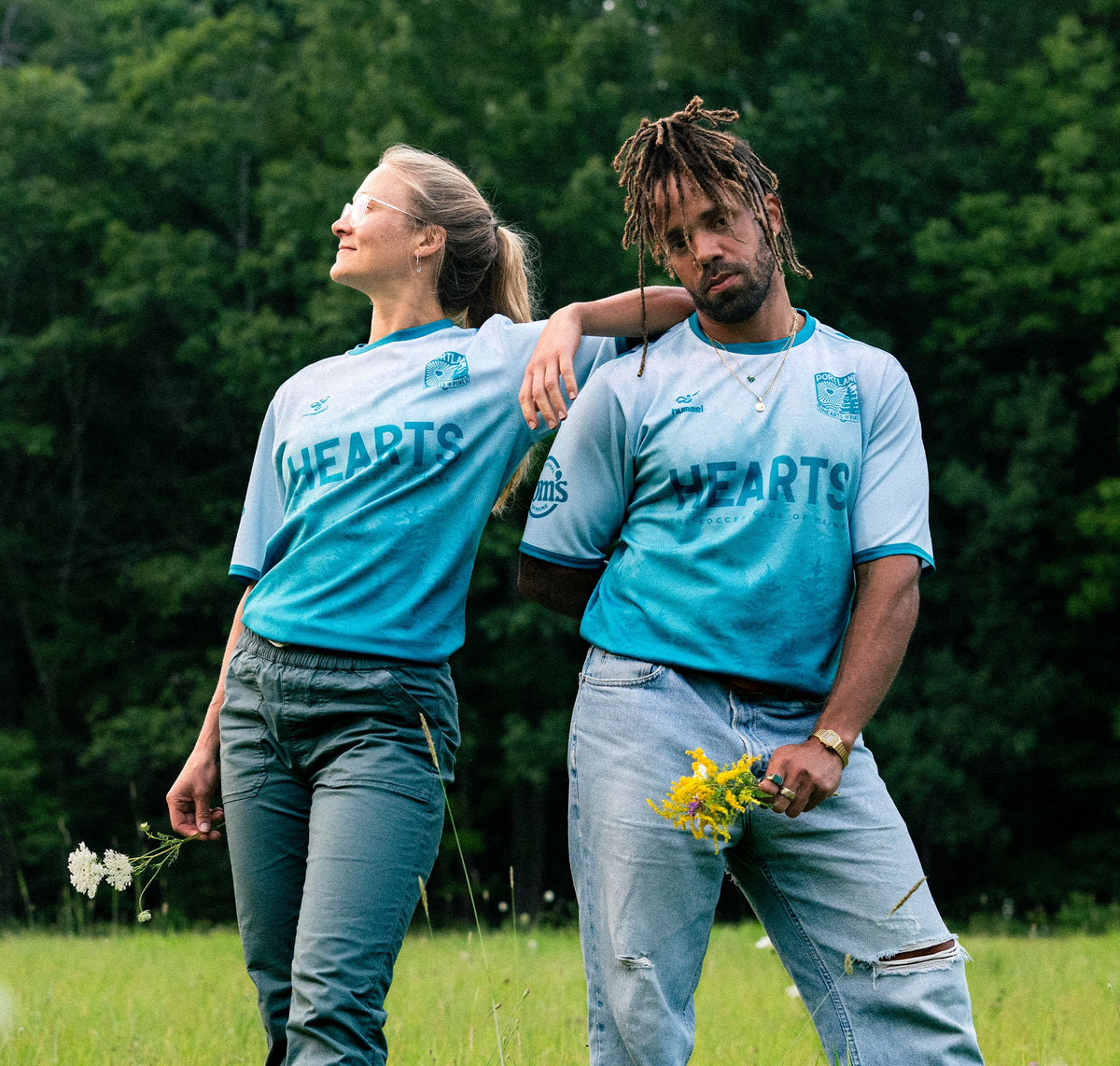 Two people in matching light blue 'HEARTS' jerseys standing in a grassy field with trees in the background.